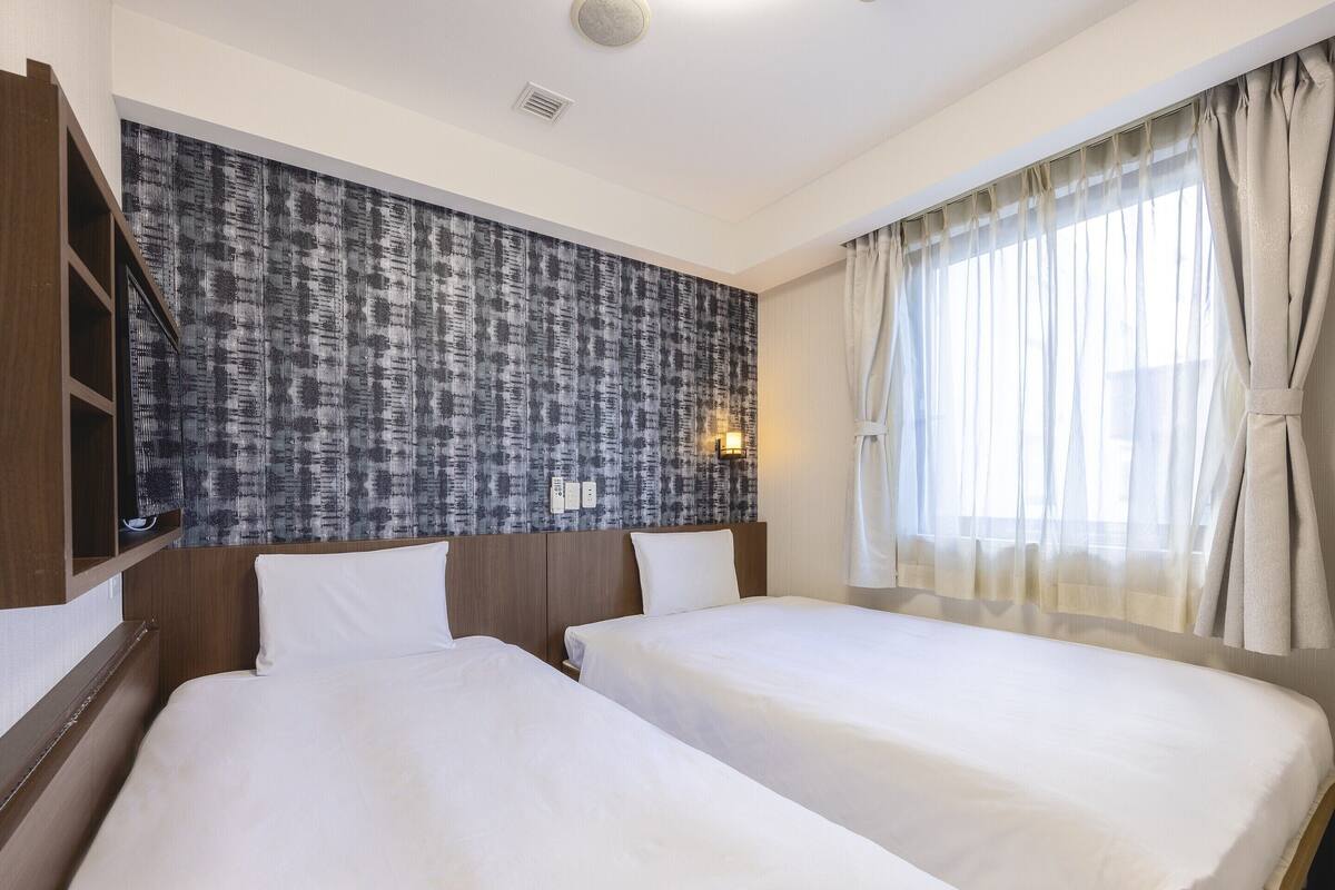 Ein Hotelzimmer mit zwei Betten, einer Wand mit Muster, einem Fenster mit Vorhängen und einer wandmontierten Lampe.