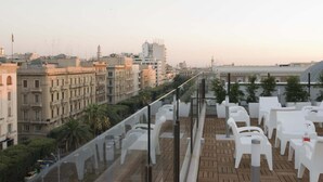 Terraza en la azotea con sillas y mesas de plástico blanco, con vistas a un paisaje urbano con edificios históricos y palmeras.