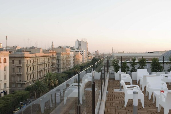 Une terrasse sur le toit, avec des chaises et des tables en plastique blanc, d’où l’on peut admirer un panorama sur la ville, avec ses bâtiments historiques et ses palmiers.