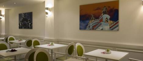 A hotel dining area with white tables and chairs, artwork on the walls, and recessed ceiling lighting.