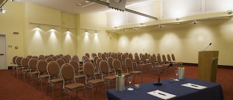 A conference room with rows of chairs, a podium, and a table with microphones and papers.