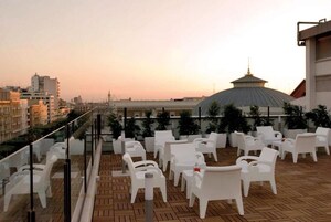 Una terraza en la azotea con sillas y mesas de plástico blanco, piso de madera y vistas a la ciudad, con un edificio de forma abovedada al fondo.
