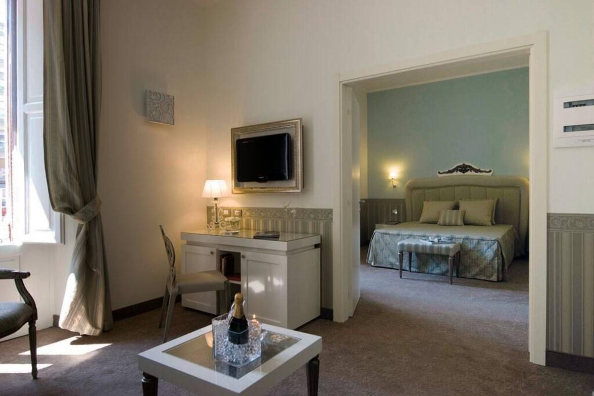 A hotel room with a bed, a desk with a television, a chair, and a small table with a bottle.