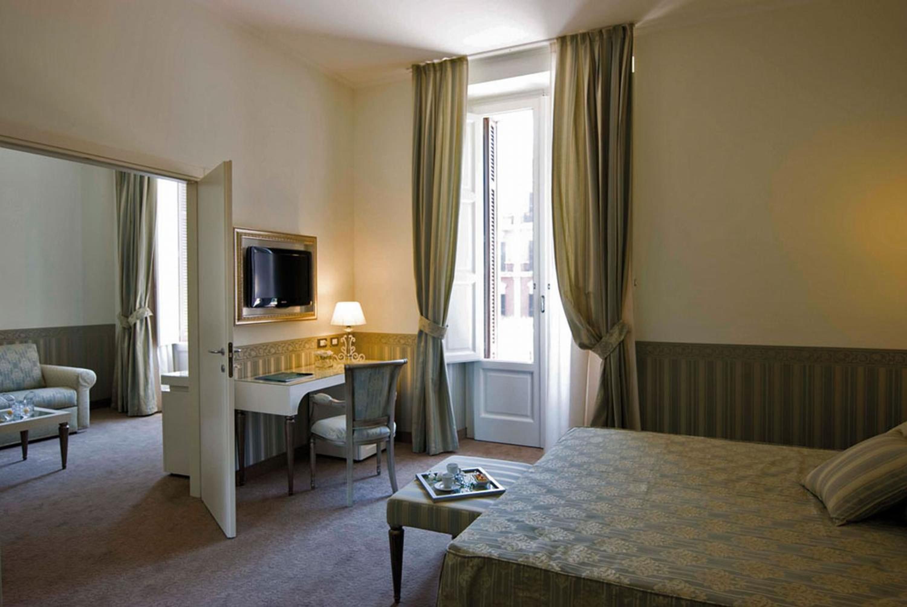 A hotel room with a bed, a desk with a chair, a television, and a window with curtains.