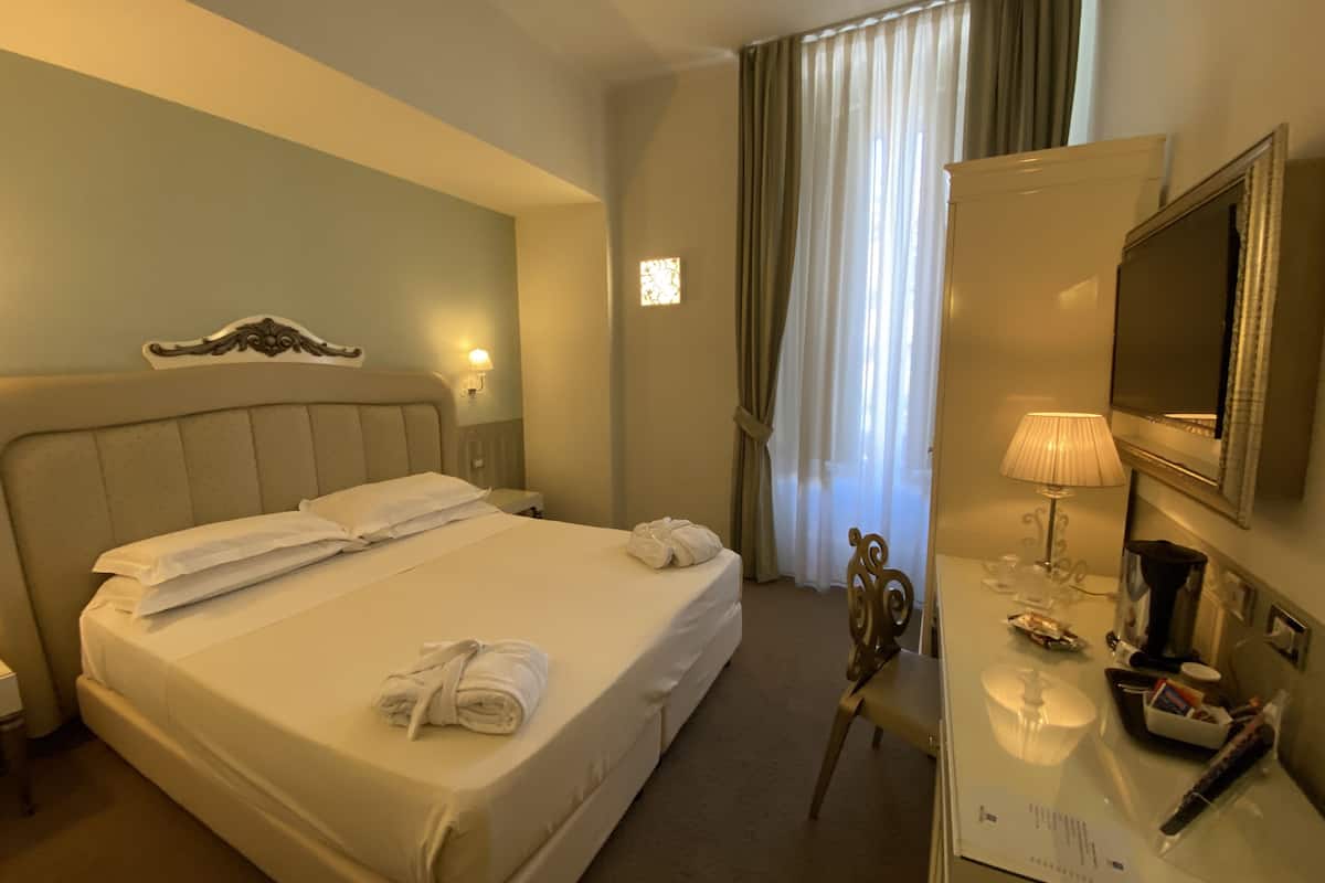 A hotel room with a large bed, a desk with a lamp, a TV, and a window with curtains.