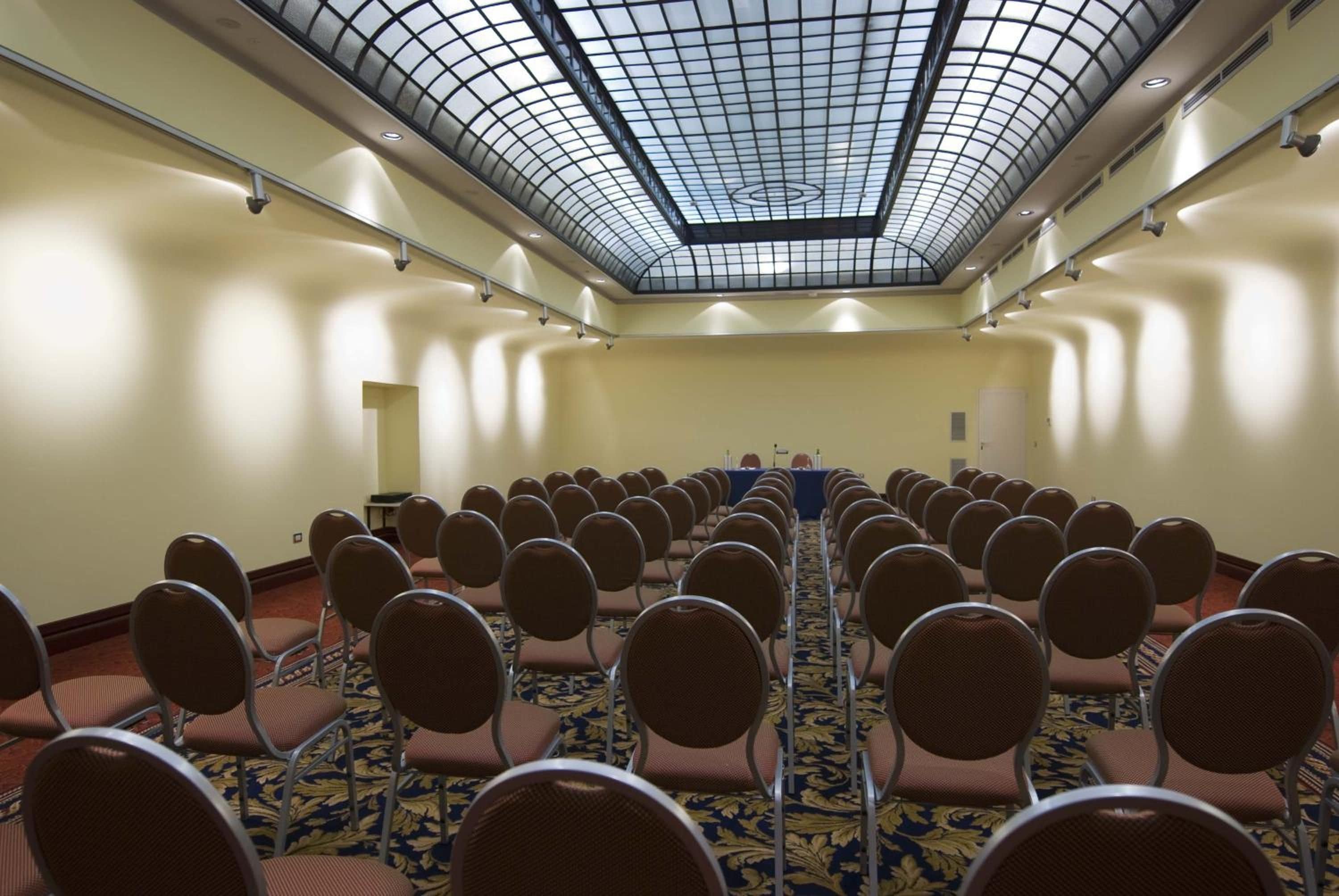Un salón de conferencias con filas de sillas frente a una larga mesa, techo de cristal y suelo alfombrado.