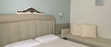 A hotel room with a large bed, a smaller sofa, a decorative headboard, a wall-mounted lamp, and a small artwork.