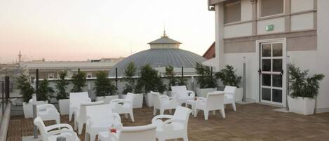 A rooftop terrace with white plastic chairs and tables, a large dome structure, and a building with an exit sign.