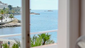 Vista costera a través de una ventana, con una playa arenosa, un muelle con palmeras y una isla distante.