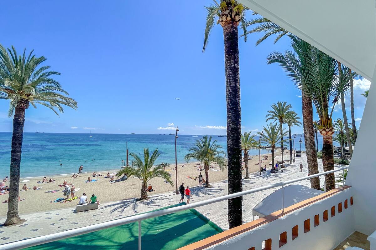 A beach view from a balcony with palm trees, a pool, and a clear blue sky.