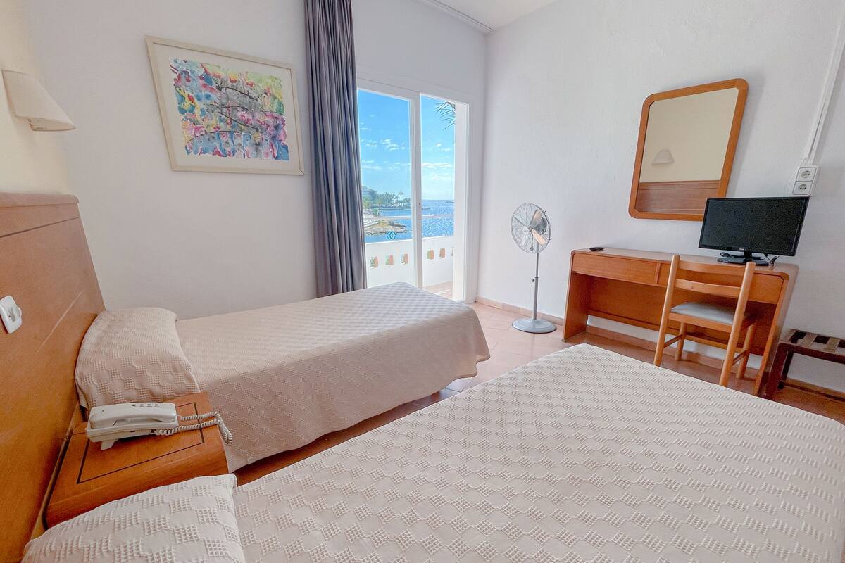 A hotel room with two beds, a desk with a computer, a standing fan, and a window with a view.