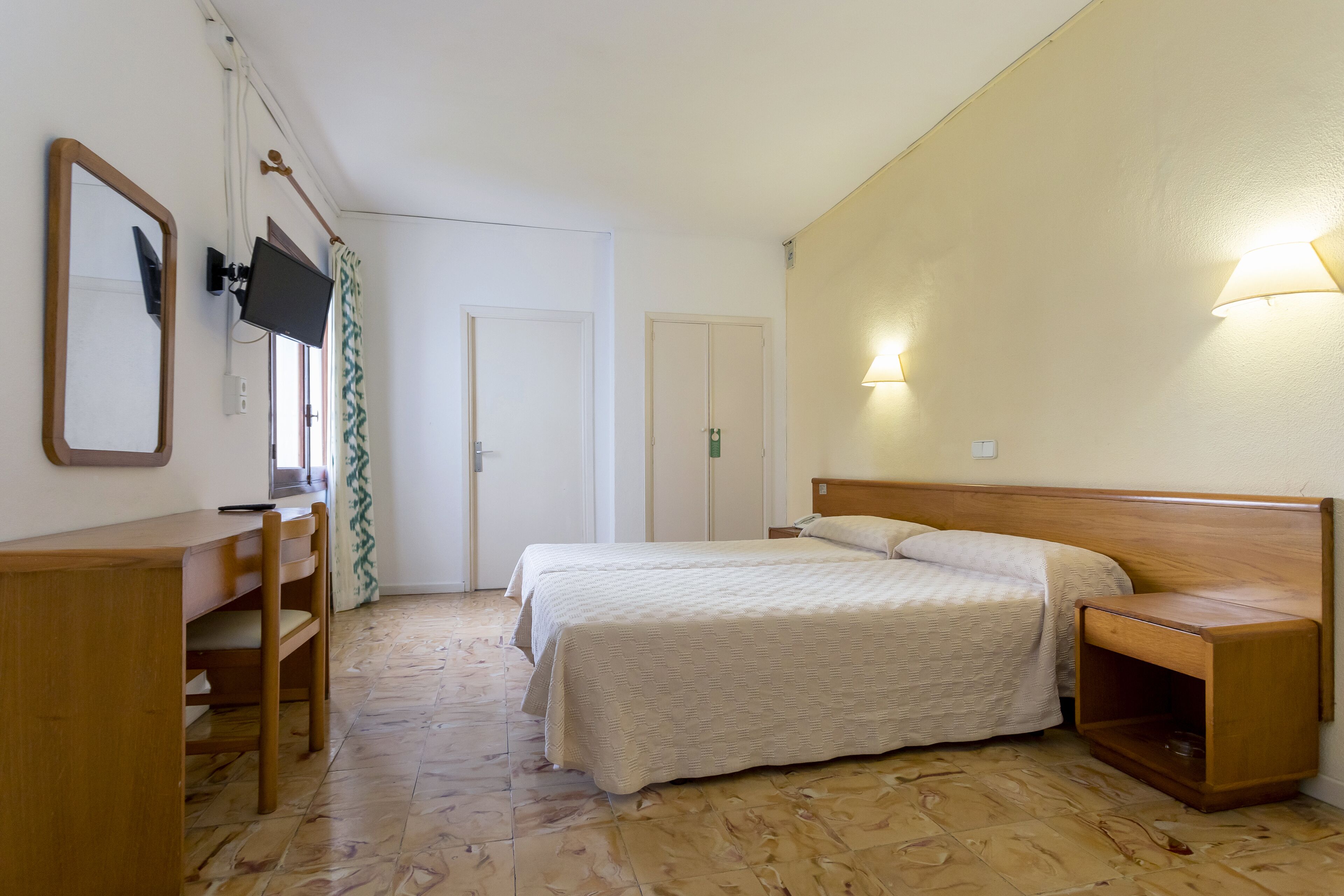 A bedroom with a bed, bedside table, wooden desk, chair, TV, and a mirror.