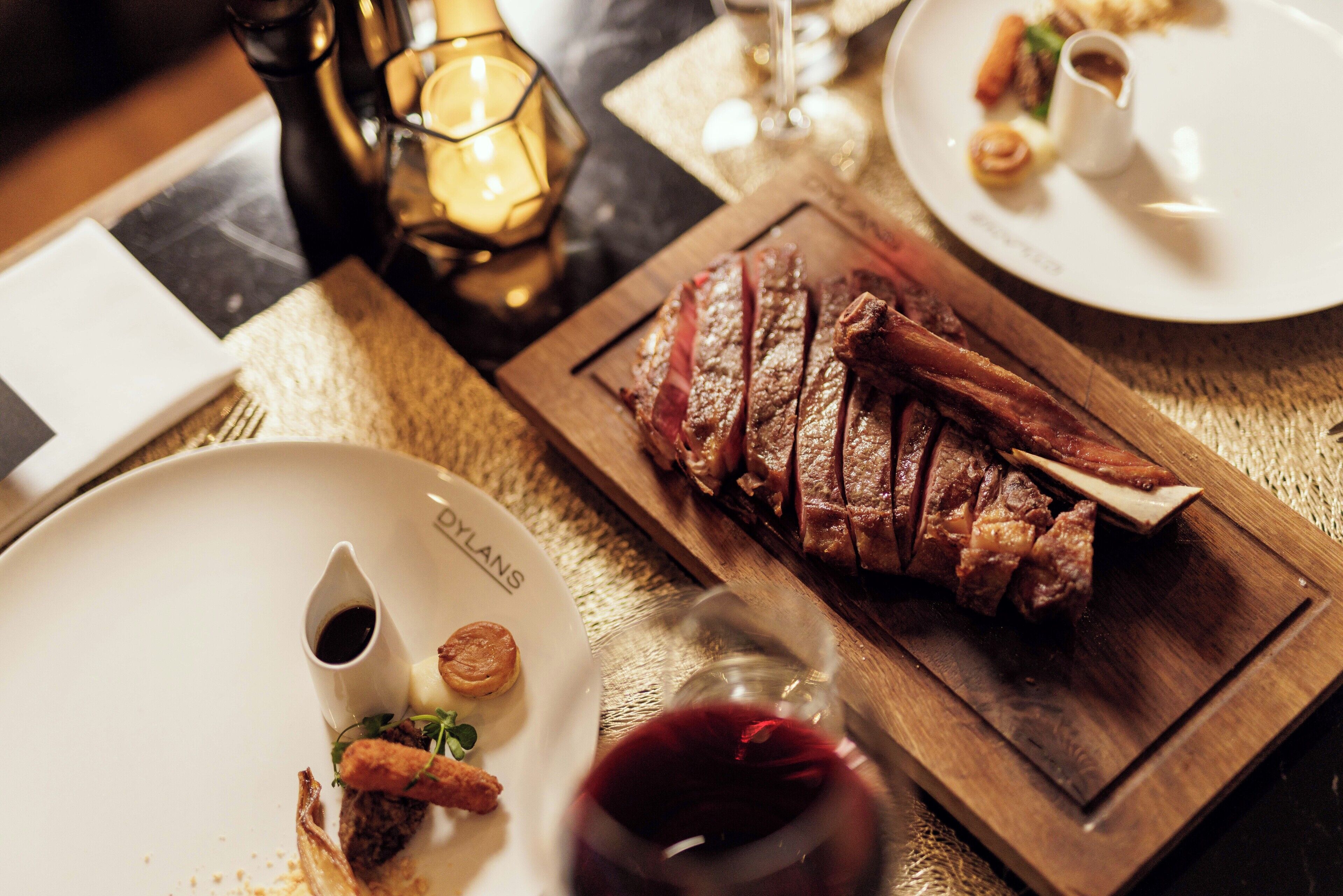 Una mesa de comedor con un plato de filete en rodajas, una copa de vino tinto y una vela.
