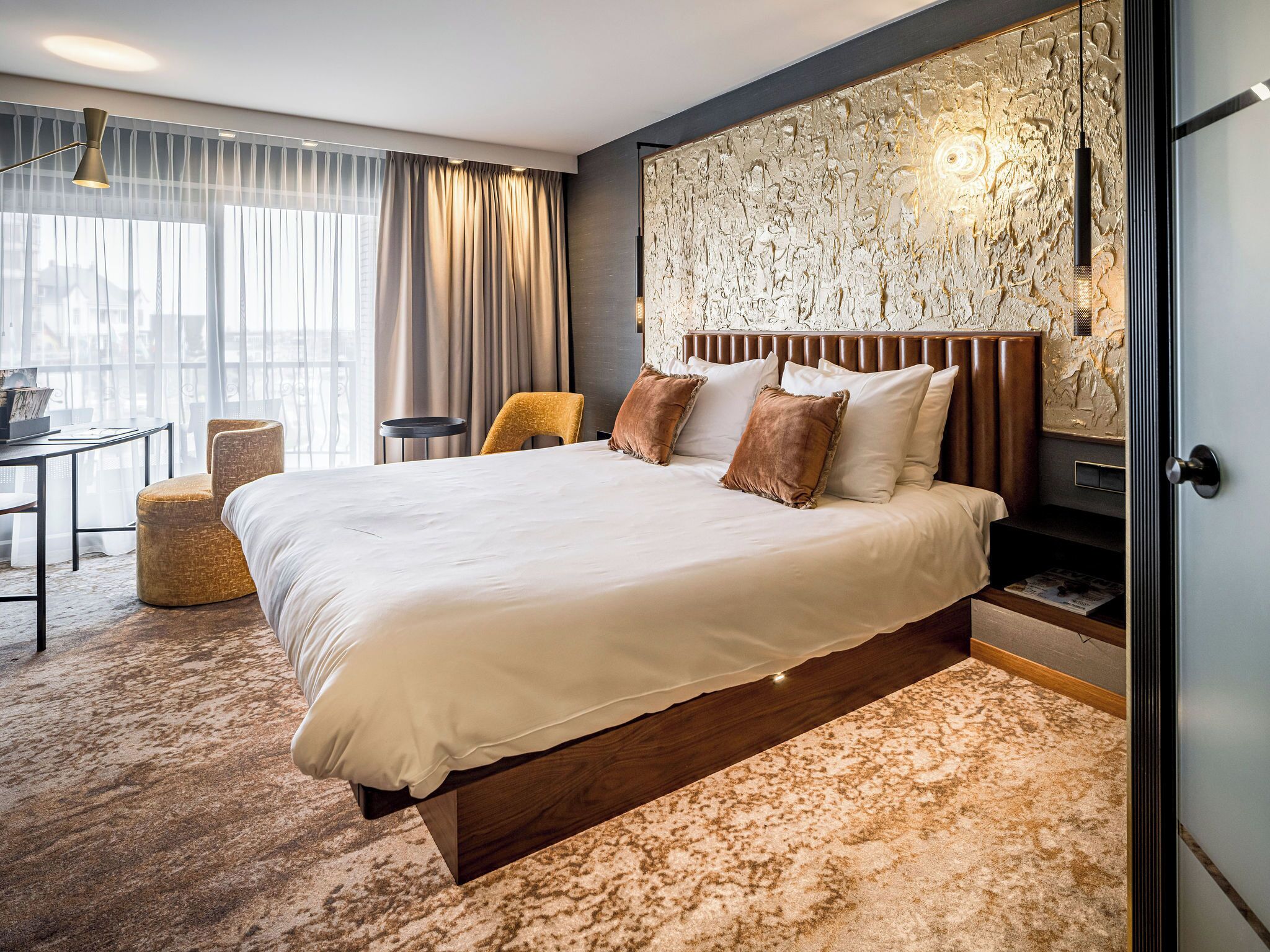A modern hotel room with a large bed, a wooden headboard, a textured wall art piece, and a view of the city through sheer curtains.