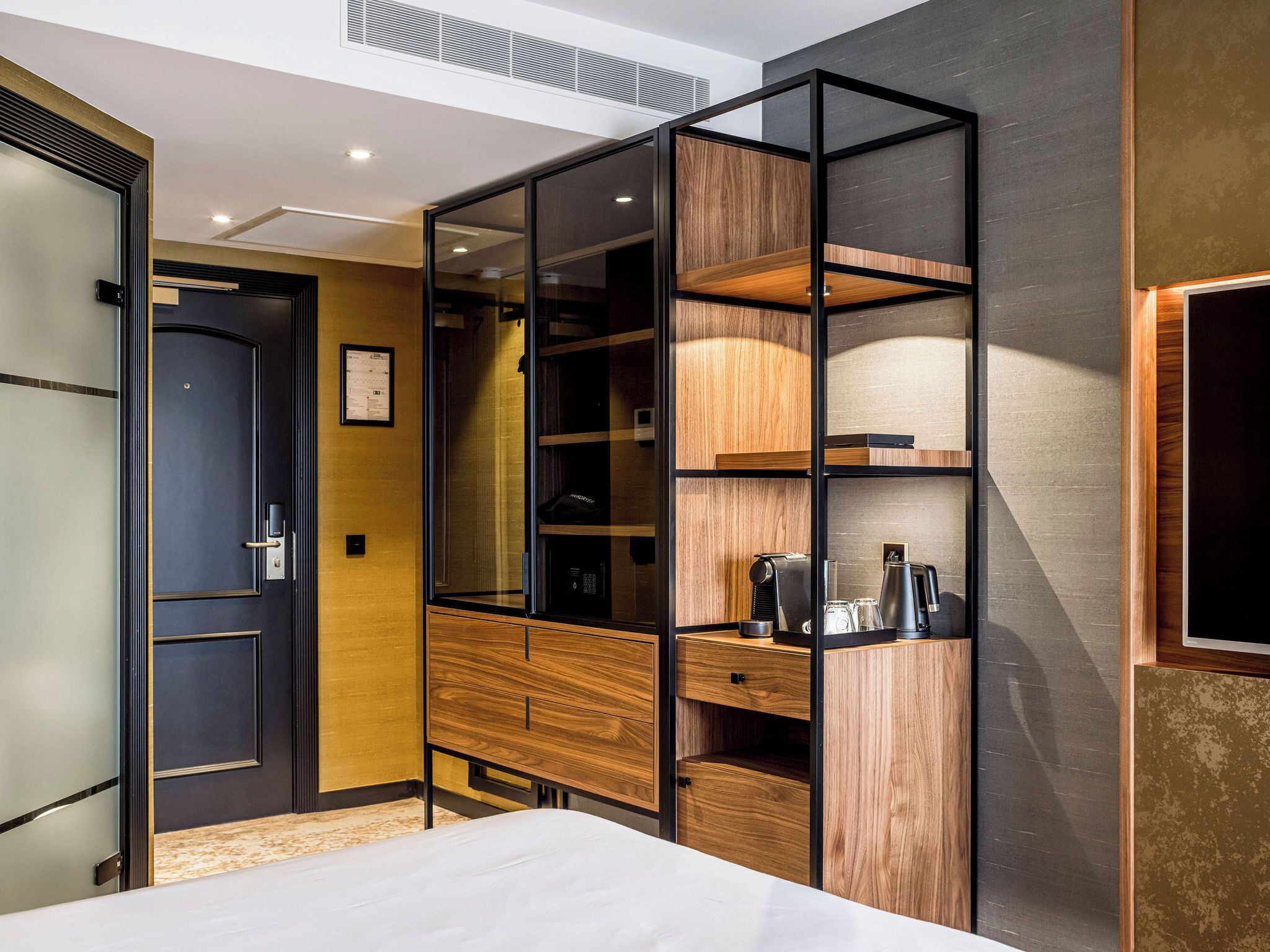 Una habitación de hotel moderna con una cama, un armario con puertas espejadas y una unidad de estanterías empotrada con una tetera.