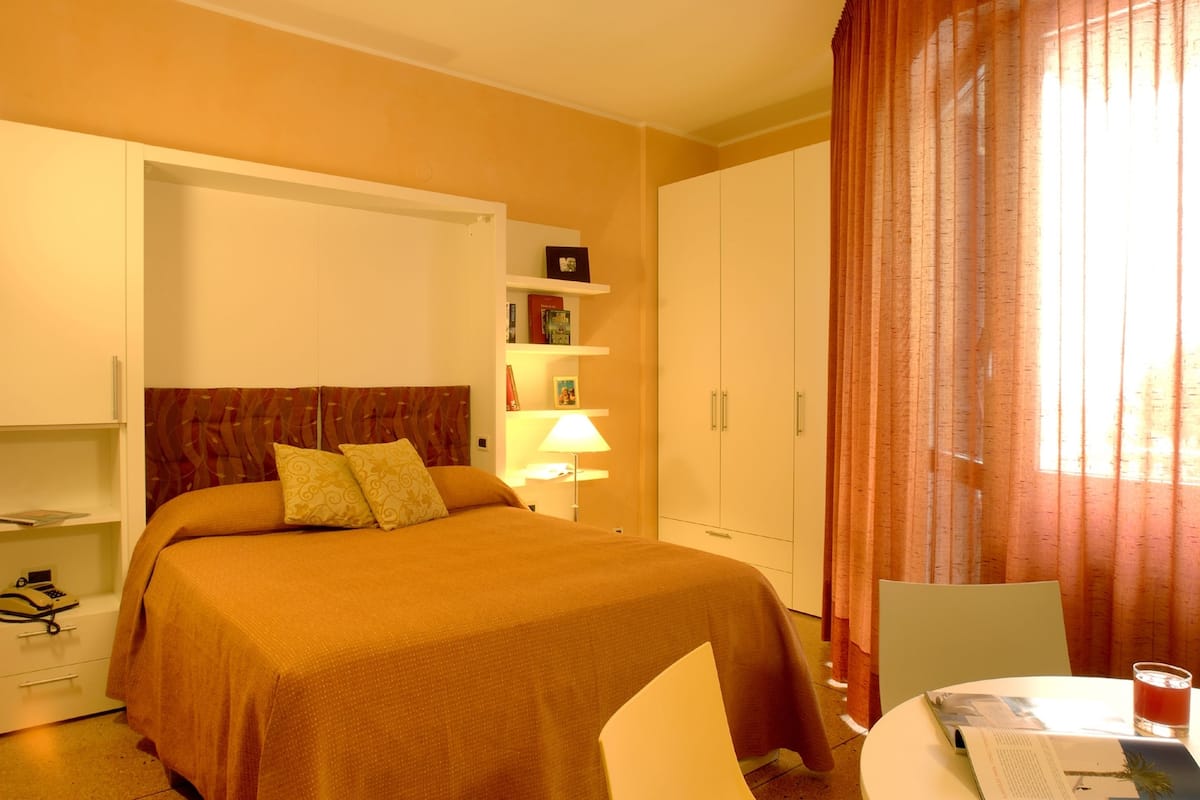Ett hotellrum med en säng, ett skrivbord med en stol, en lampa, en bokhylla och en garderob.