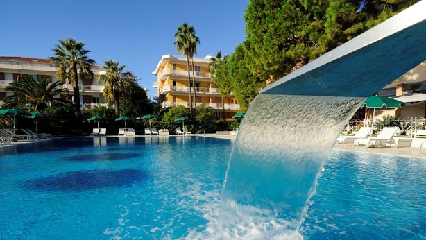 A swimming pool with a water slide, surrounded by lounge chairs and palm trees, with a hotel building in the background. - Residence Oliveto (Ceriale)