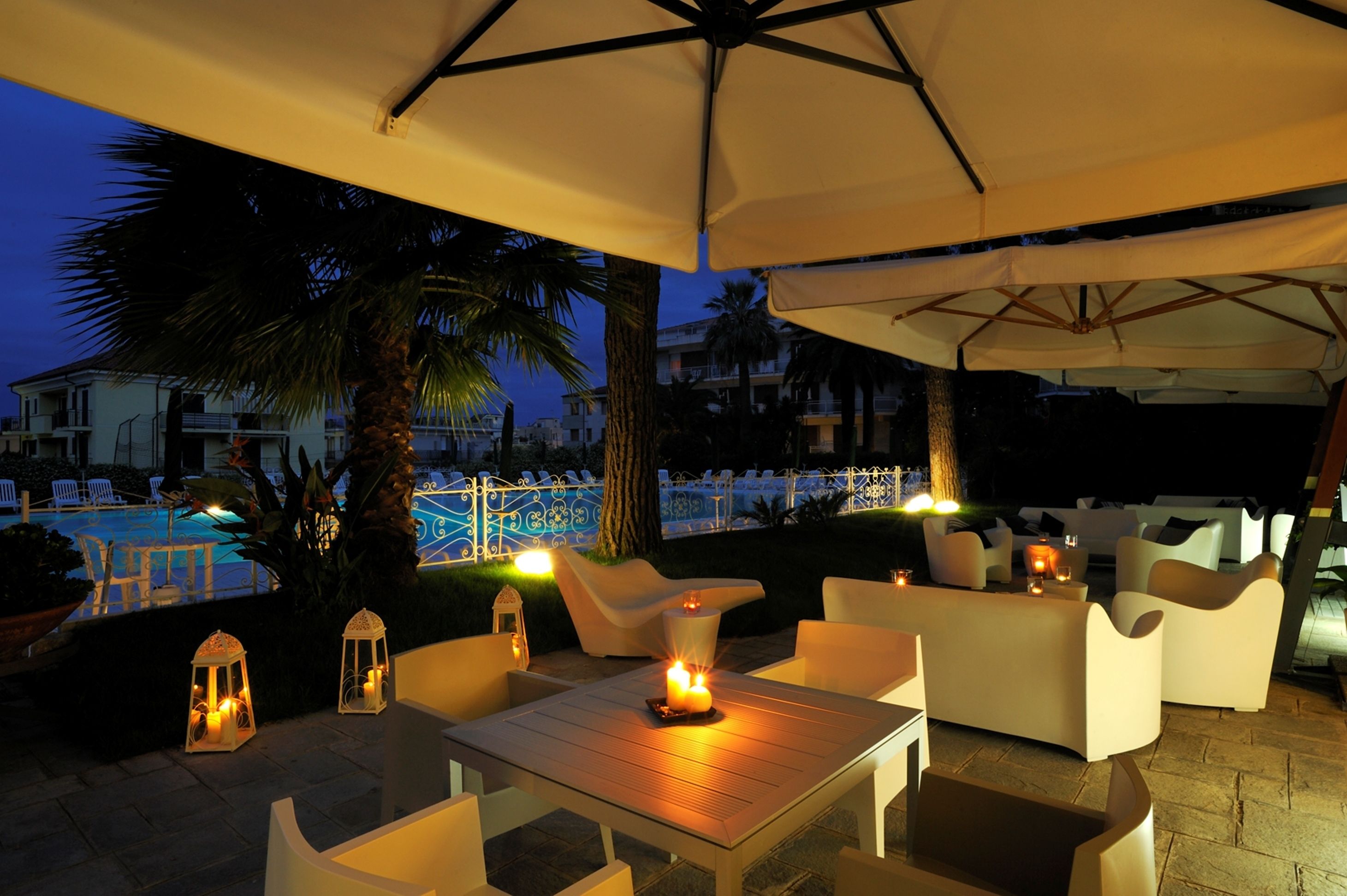 Un área de estar al aire libre con muebles blancos y una mesa con velas, con vistas a una piscina y palmeras.