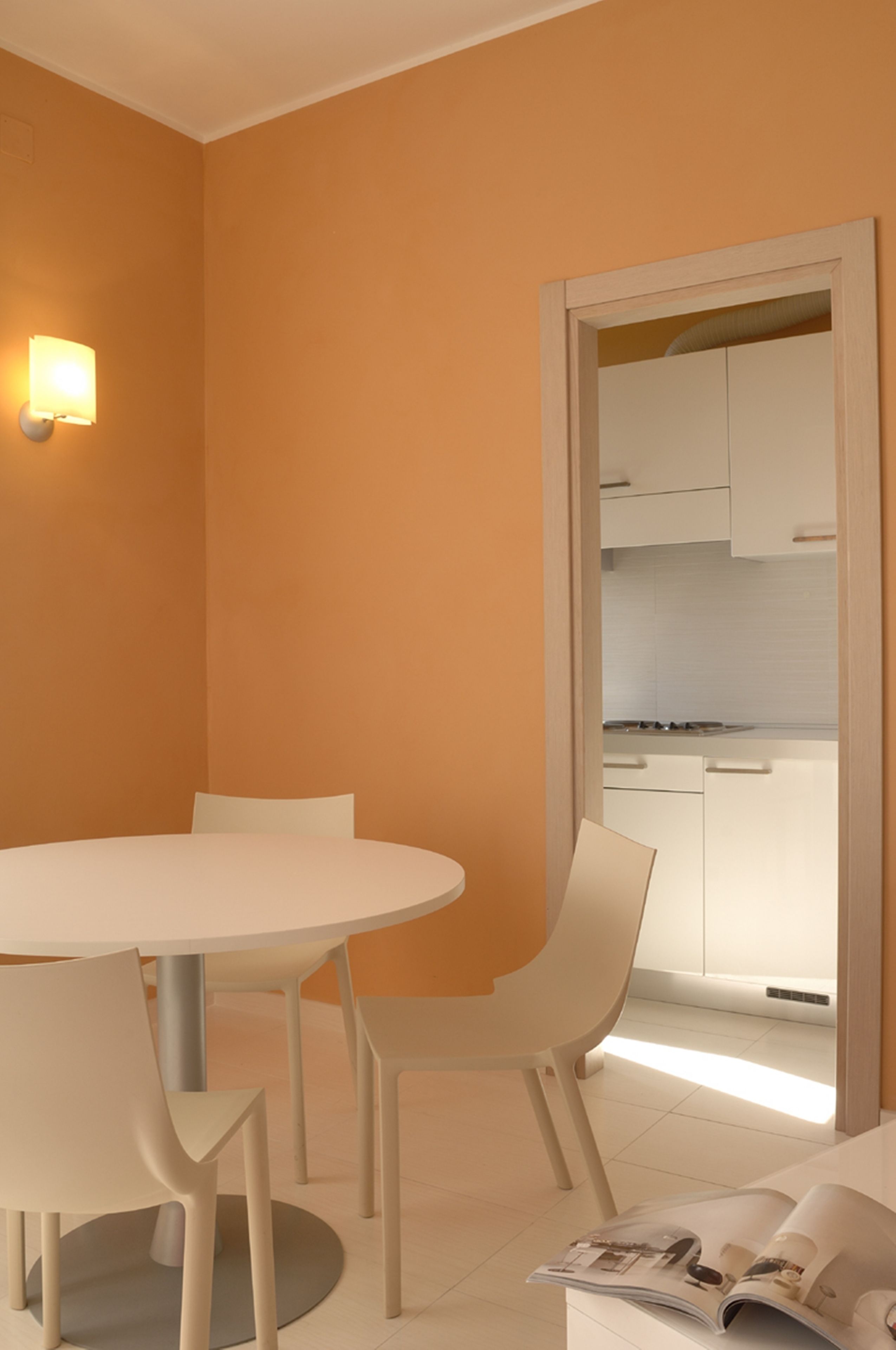 Un espace repas moderne avec une table ronde et des chaises, un luminaire fixé au mur, et une vue sur une cuisine aux armoires blanches.