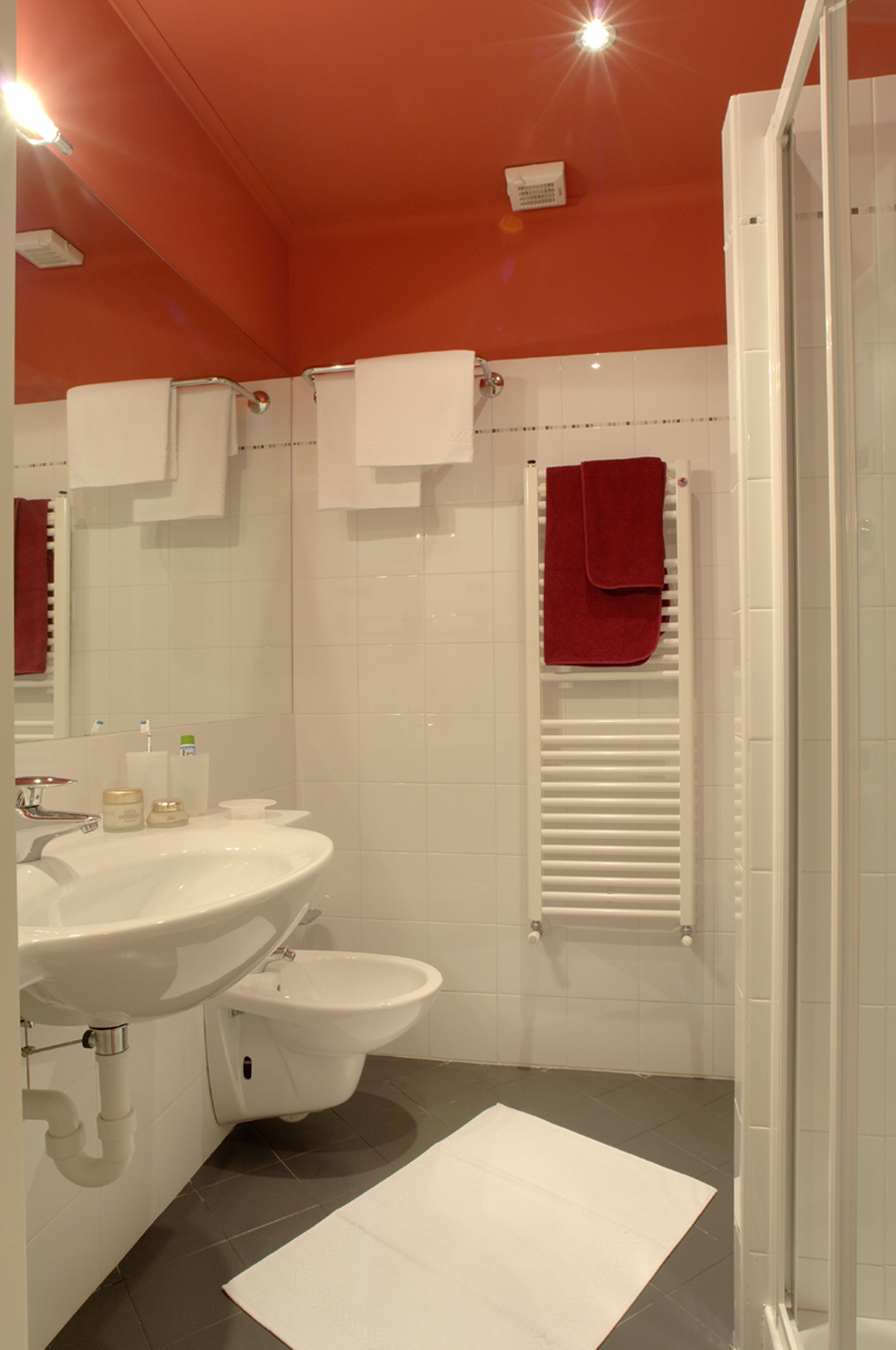 Une salle de bain avec un lavabo blanc, un bidet et une douche. Des serviettes sont accrochées au mur et une serviette rouge est posée sur la tringle de la douche.