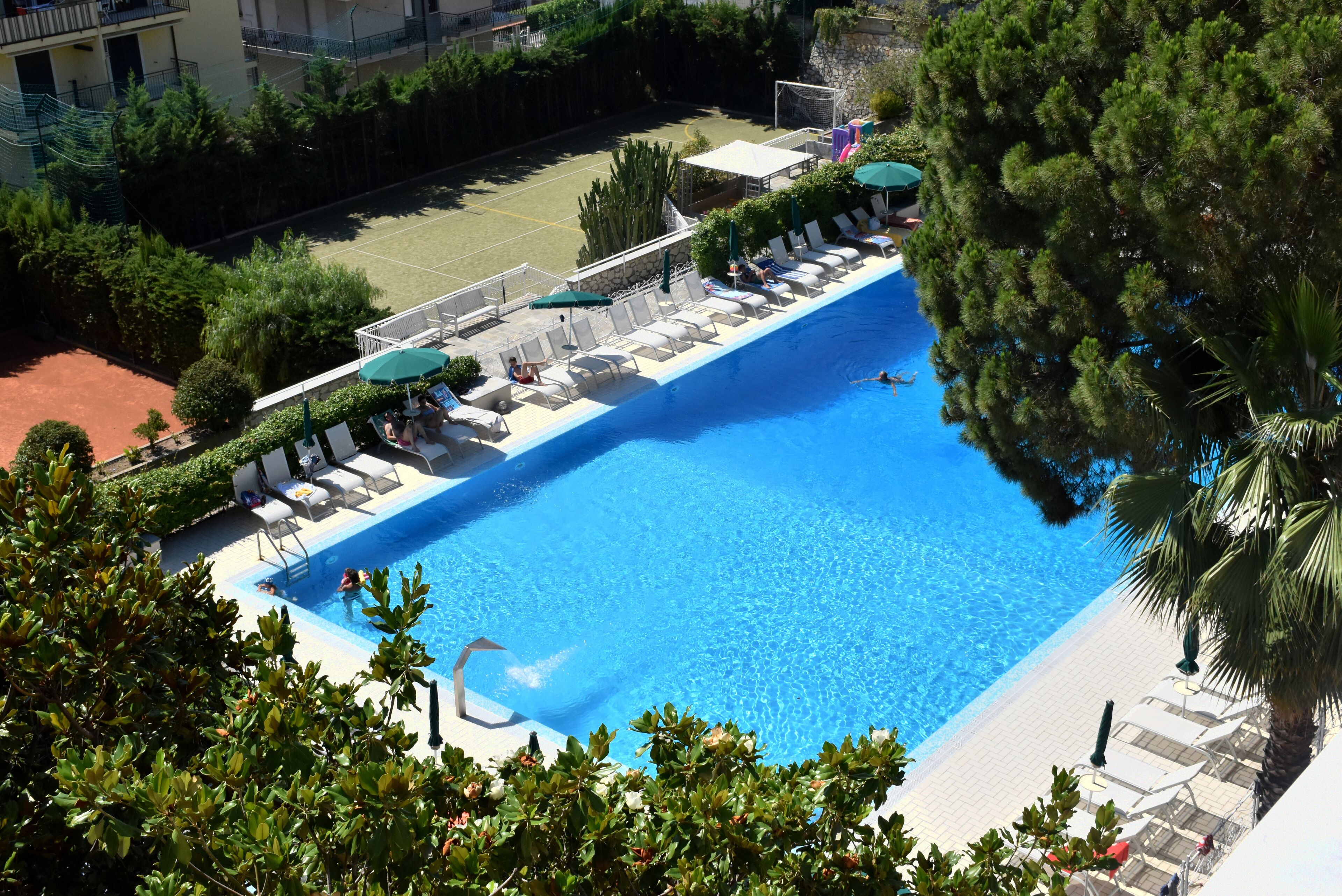 A swimming pool with lounge chairs and umbrellas, surrounded by greenery and a tennis court.