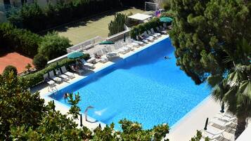 A swimming pool with lounge chairs and umbrellas, surrounded by greenery and a tennis court.
