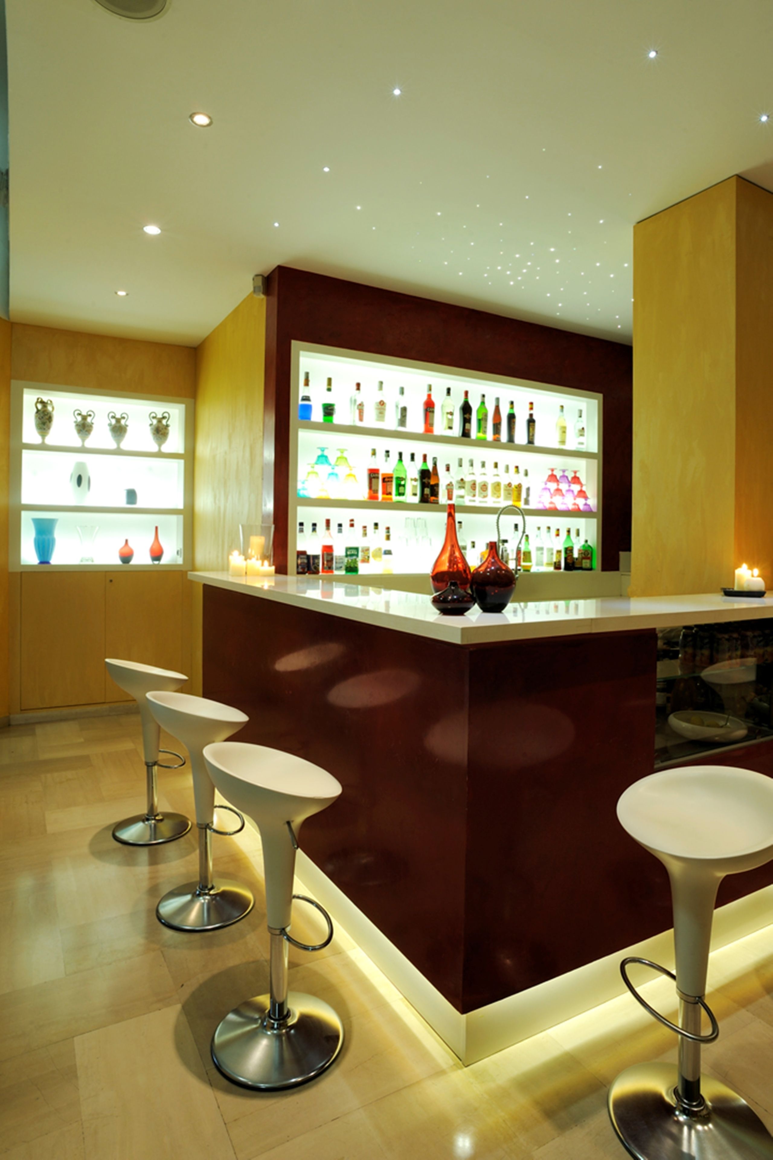 Zona de bar con una isla central, taburetes y estantes llenos de botellas y vasos.