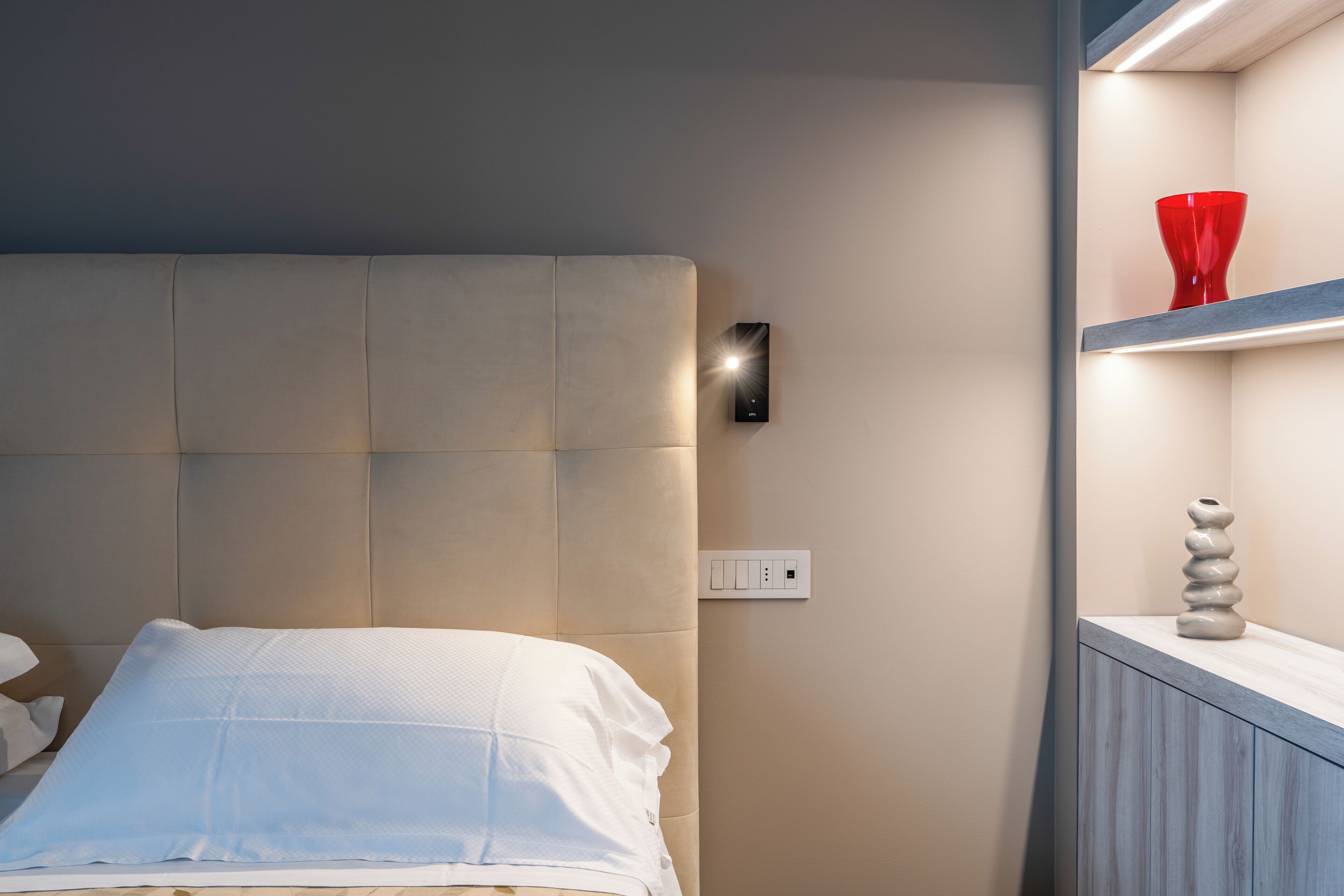 Une chambre moderne avec une tête de lit rembourrée, une table de chevet ornée d’un vase rouge et un lampadaire fixé au mur.