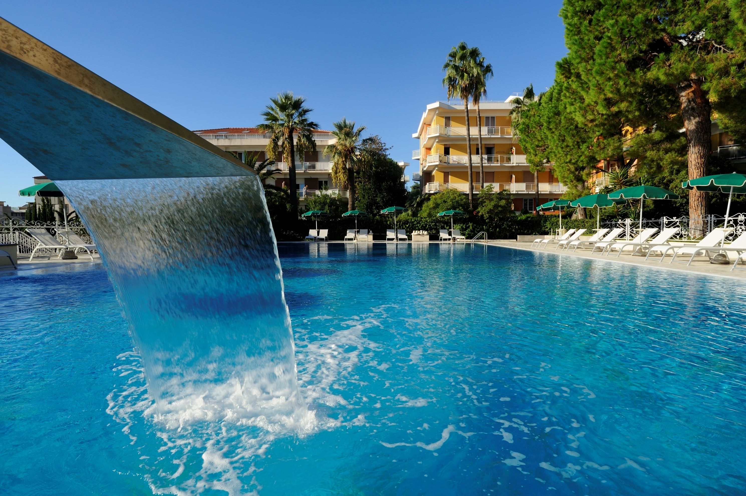 Una piscina con cascada, sillas de descanso, palmeras y un edificio de hotel al fondo.