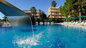 Una piscina con cascada, sillas de descanso, palmeras y un edificio de hotel al fondo.