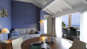 Una habitación con pared azul, un sofá con cojines, una mesa con una revista, una lámpara, un teléfono y vistas al océano a través de amplios ventanales.