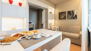 A modern hotel room with a breakfast setup on a white table, a beige sofa, and artwork on the wall. - Residence Oliveto (Ceriale)