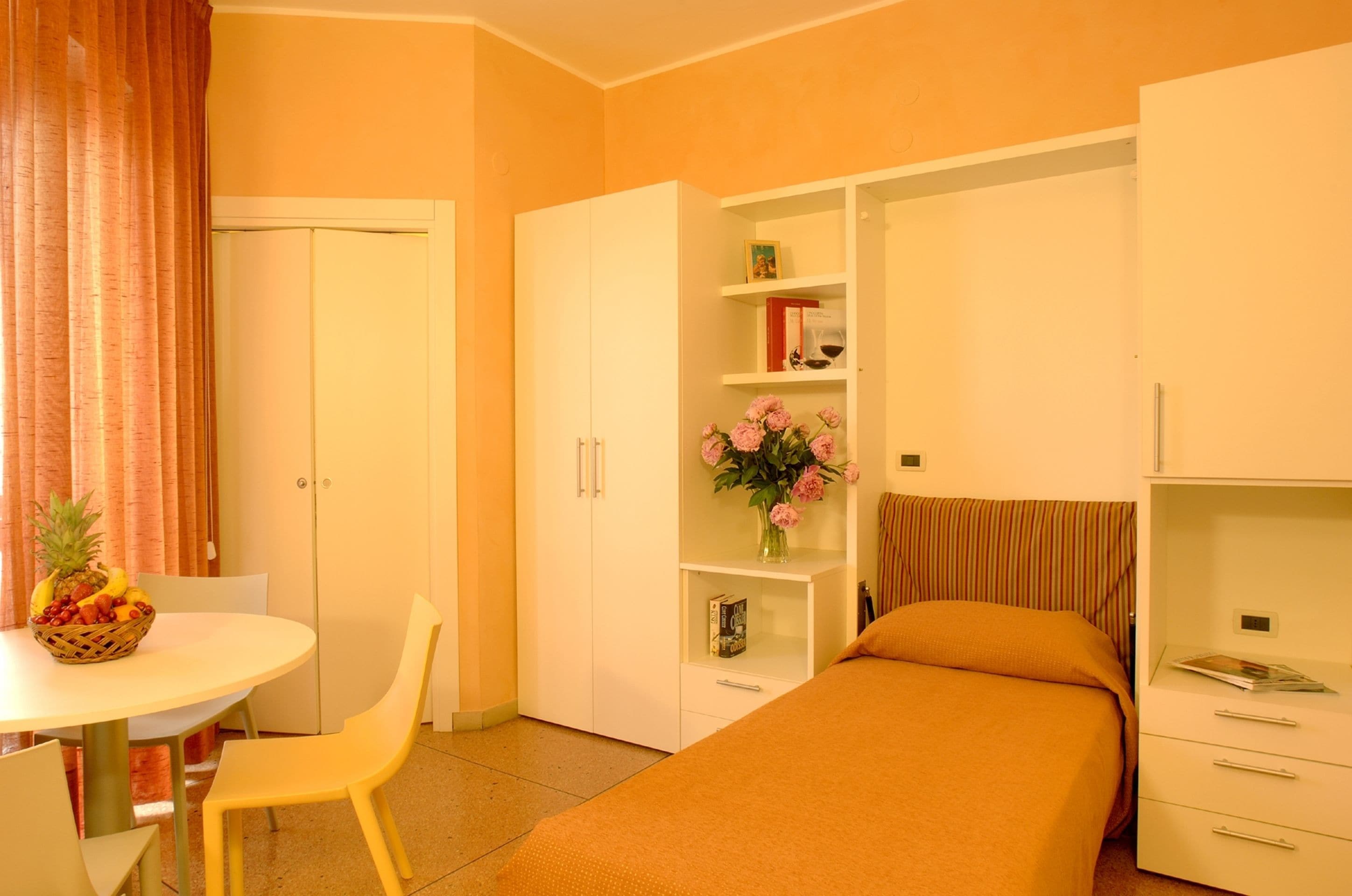 A hotel room with a bed, a desk with a chair, a wardrobe, and a small shelf with flowers.