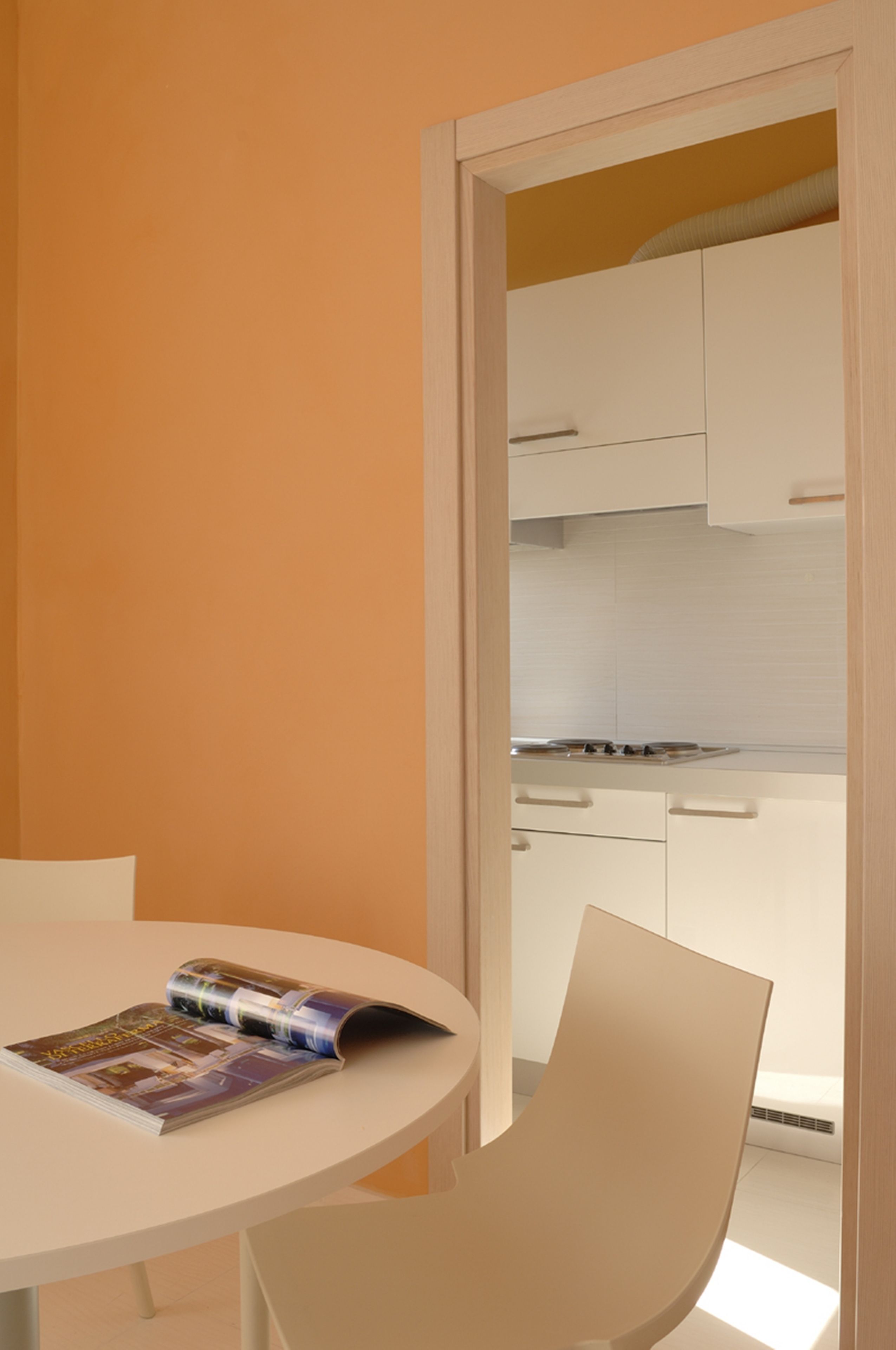 A room with a white table and chair, a magazine on the table, and a kitchen visible through an open door.