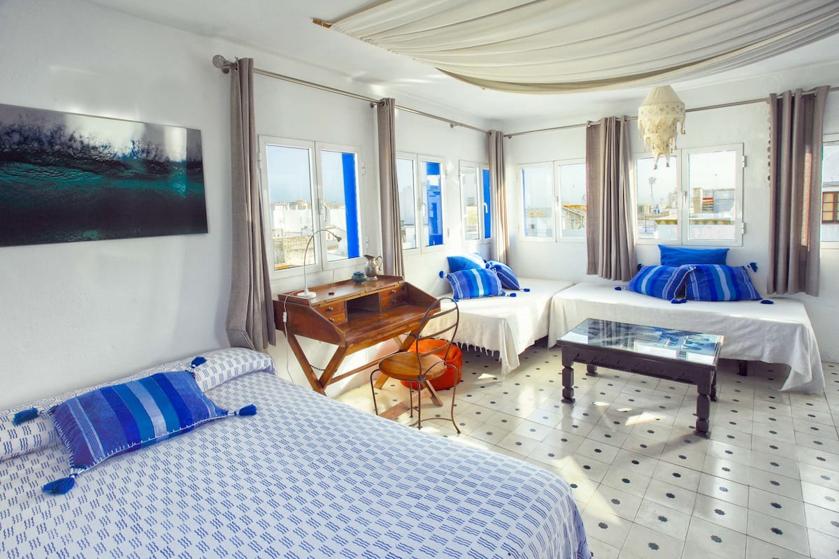 A hotel room with two beds, a desk, and a chair. There are curtains on the windows, and a painting on the wall.