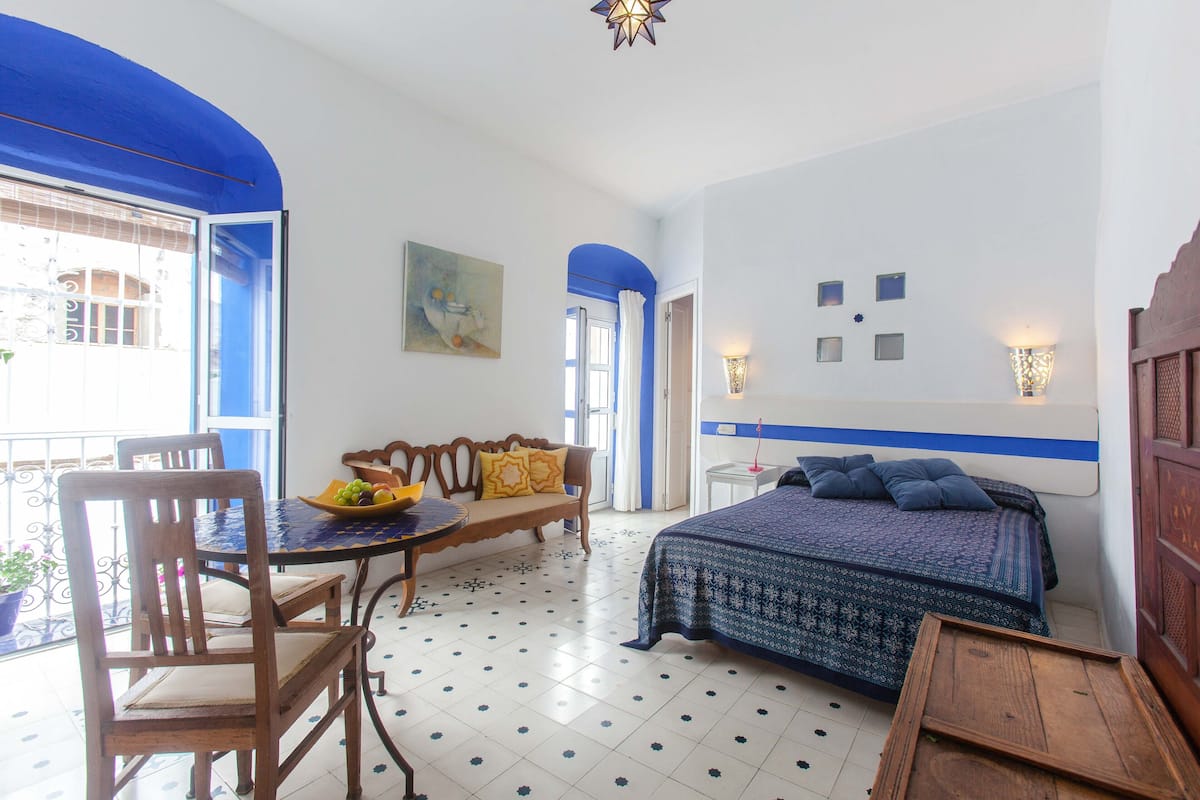 A room with a bed, a sofa, a dining table, and a chair. There is a window with blue curtains, a wall with a painting, and a wall-mounted light fixture.