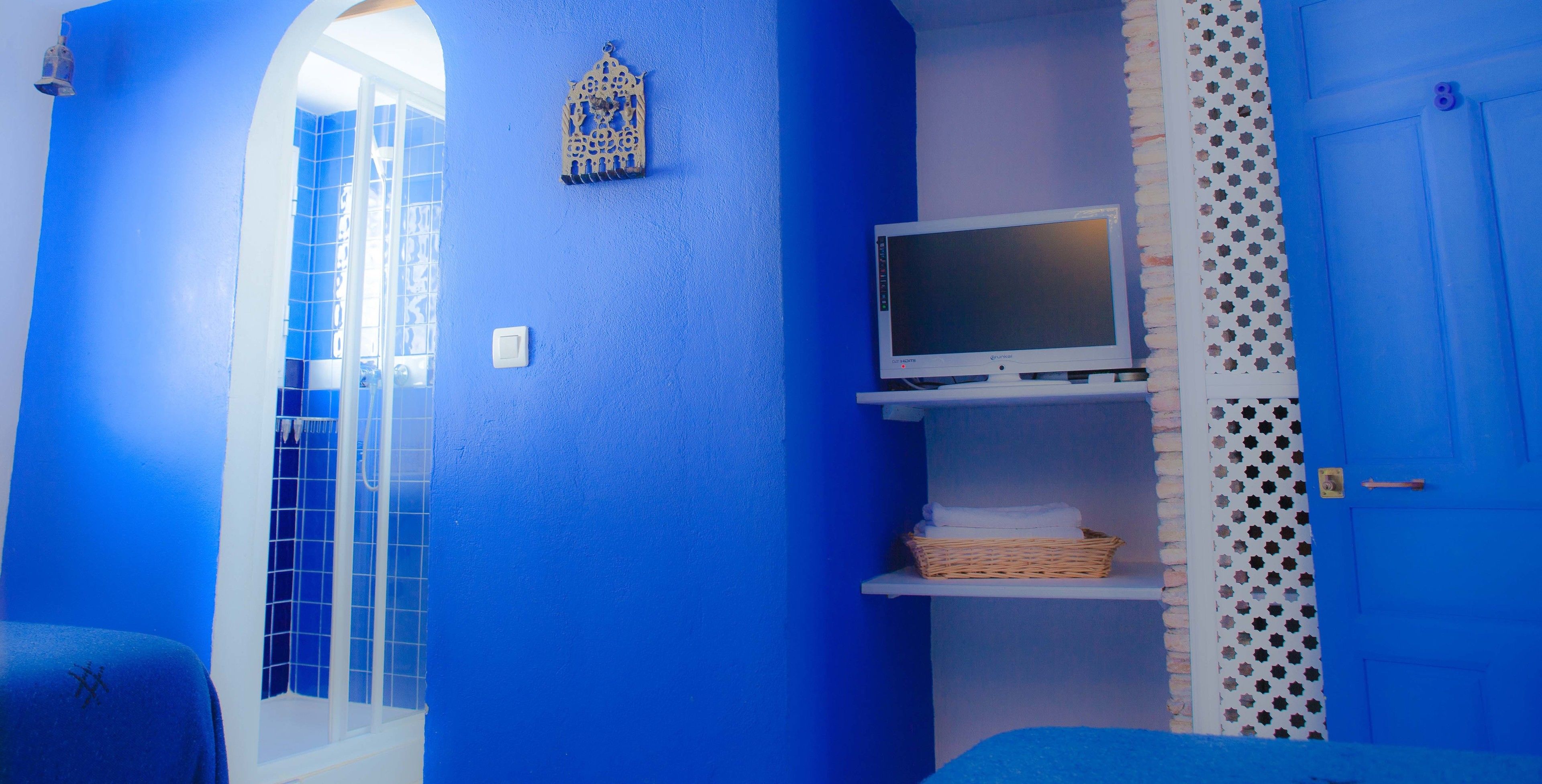 A room with a blue wall, a TV on a shelf, a door with a number, and a window with a grid pattern.