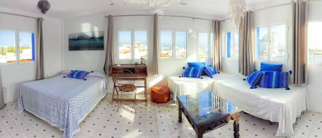 A hotel room with two beds, a desk, a chair, a small table, and large windows offering a city view.