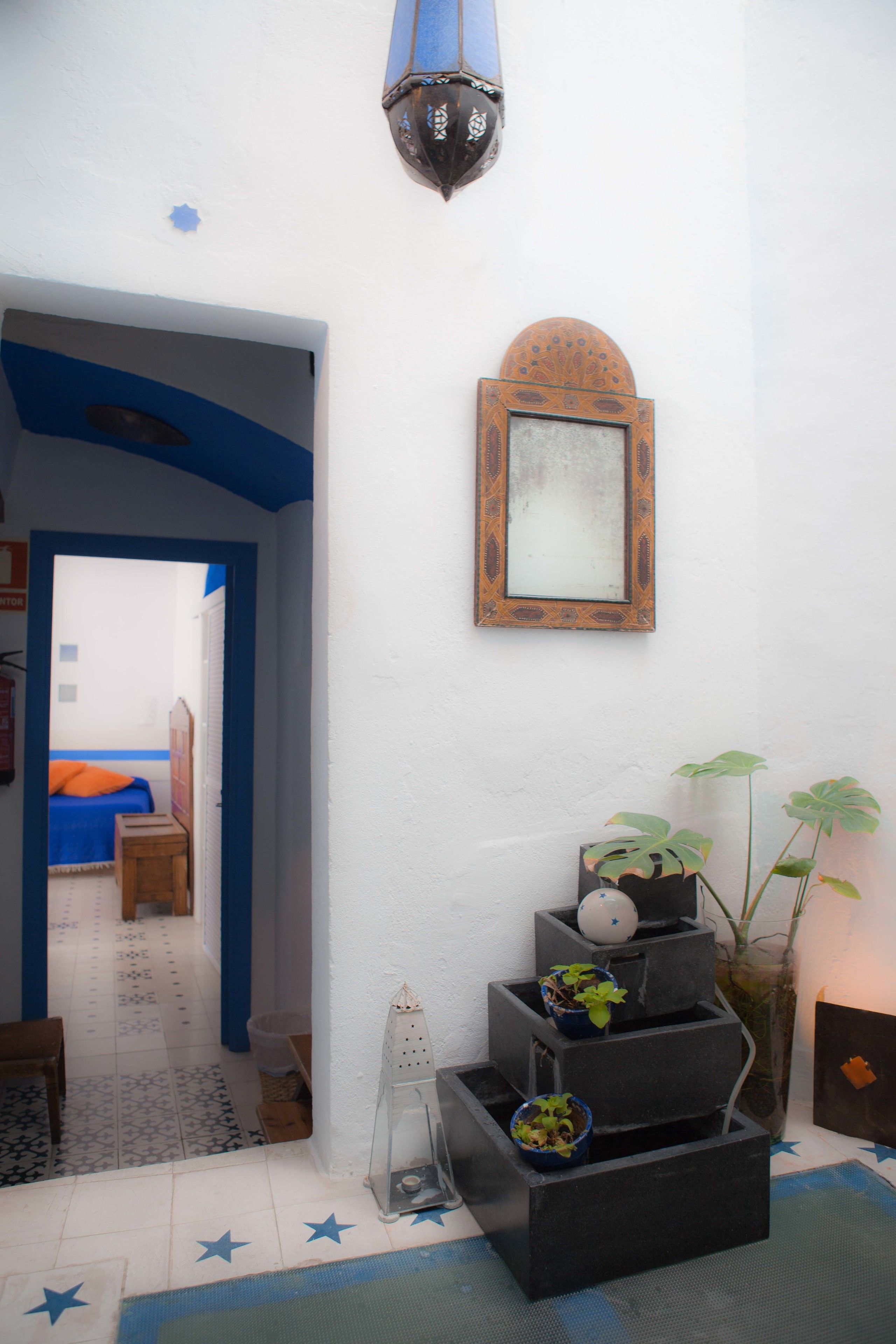 A hallway with a blue ceiling, a wooden bed with an orange bedspread, a mirror on the wall, and a staircase with potted plants.