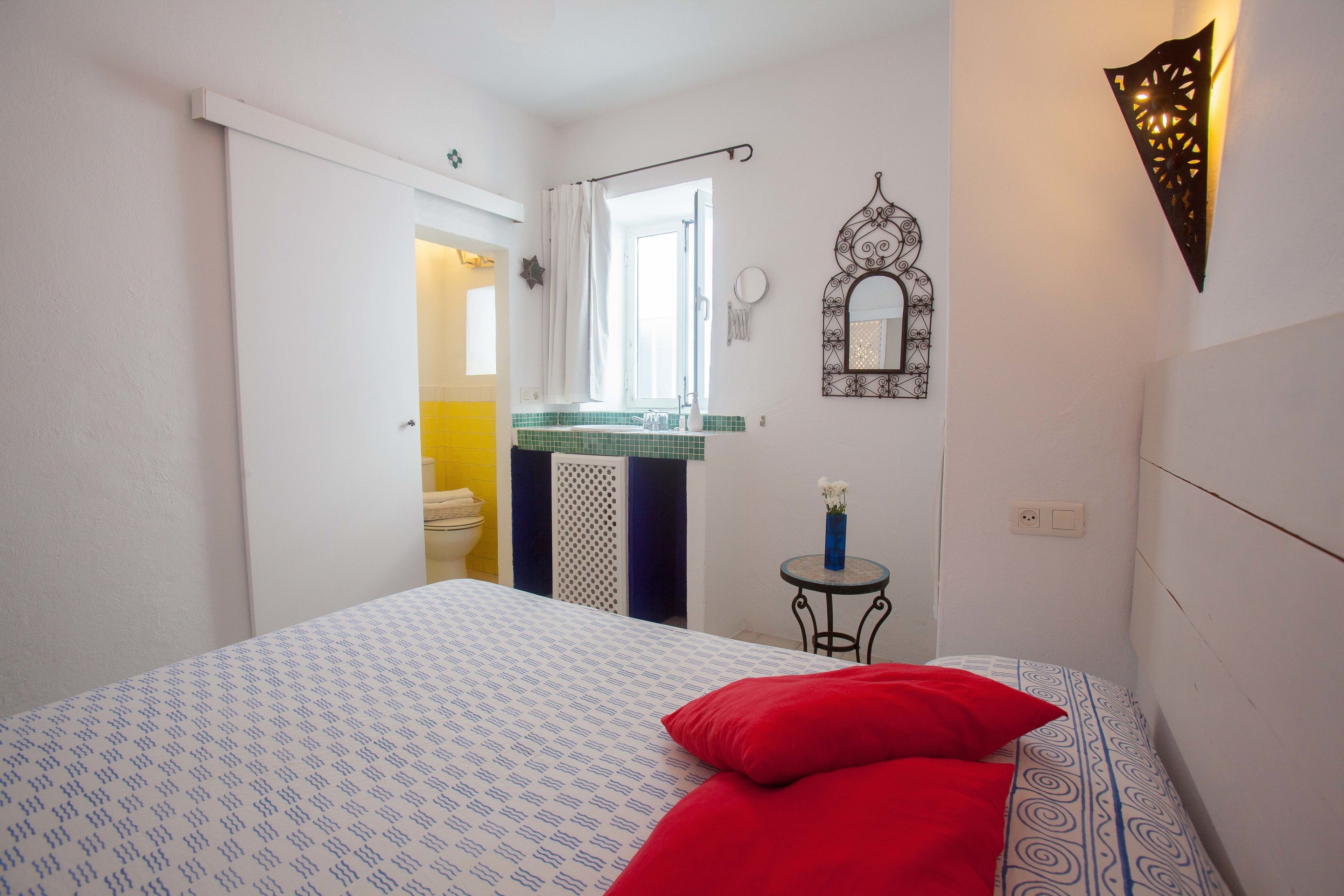 A bedroom with a bed, a nightstand, a mirror, and a bathroom visible through an open door.