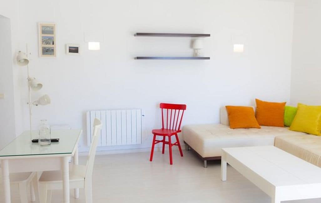 A room with a white table, a red chair, a white sofa with colorful cushions, and a white radiator.