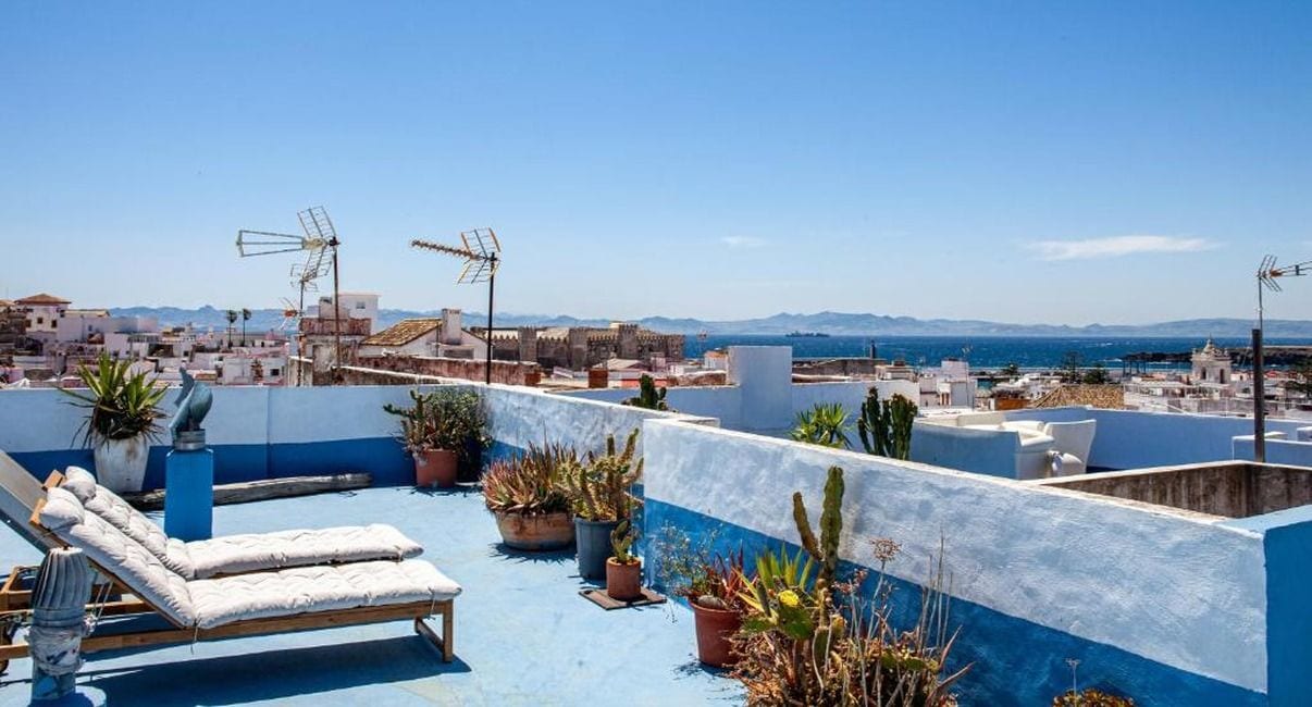 Una terraza en la azotea con una silla de descanso, plantas en macetas y vistas a un pueblo costero.