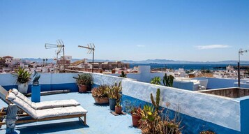 Una terraza en la azotea con una silla de descanso, plantas en macetas y vistas a un pueblo costero.