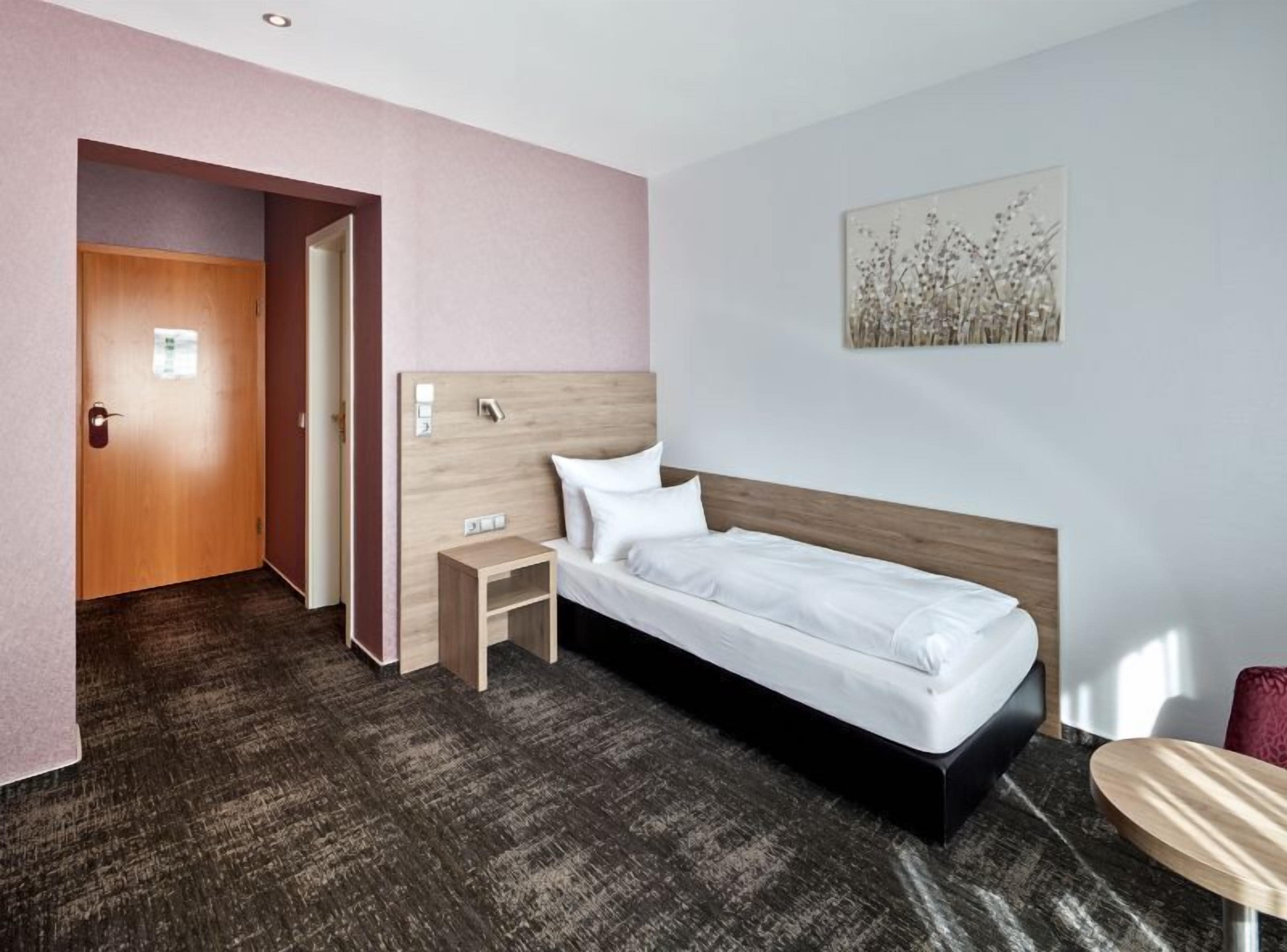 A hotel room with a single bed, a bedside table, a small nightstand, a door, and a wall-mounted artwork.