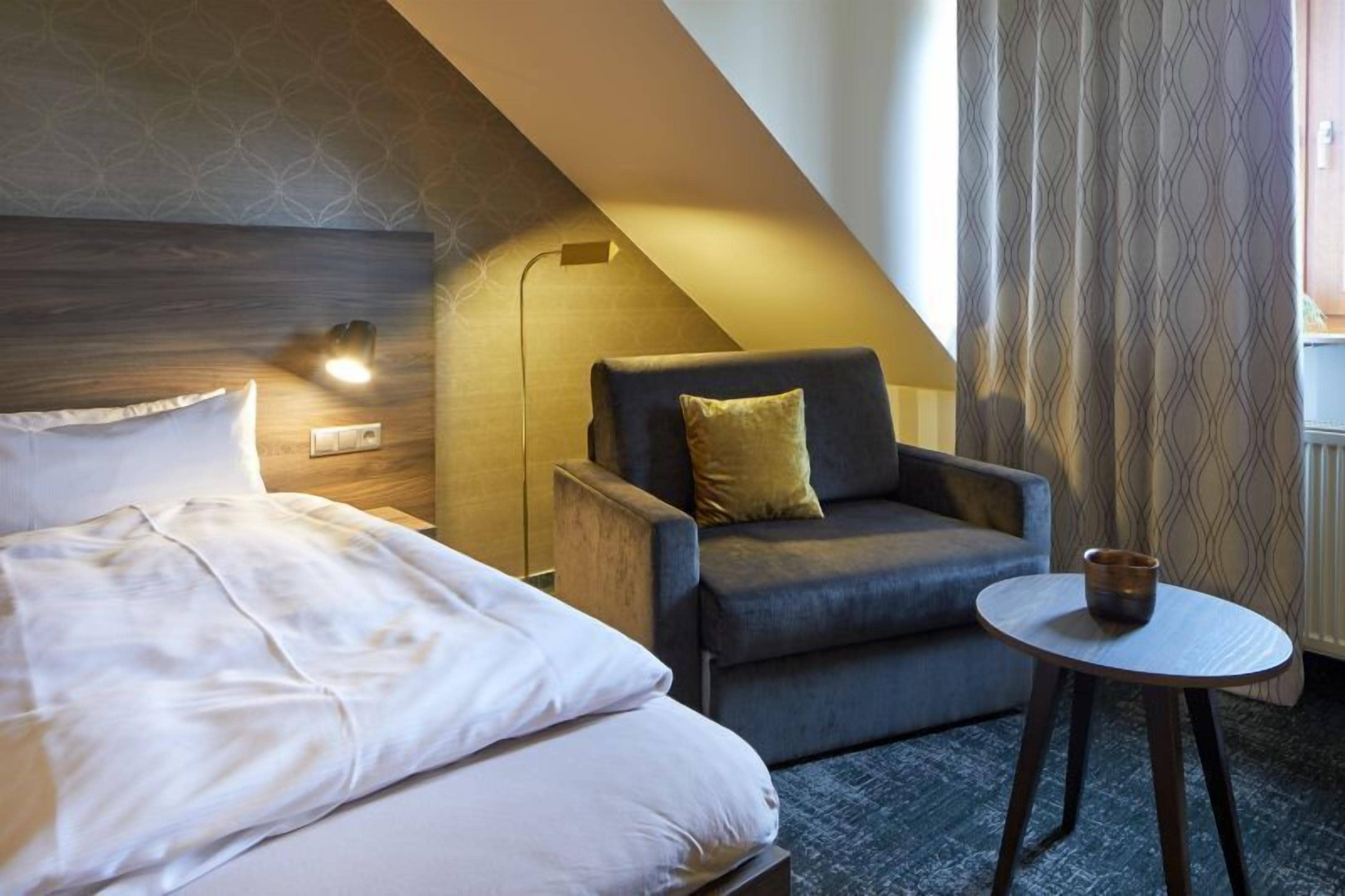 Ein Hotelzimmer mit einem Bett, einem grauen Sessel mit gelbem Kissen, einem kleinen runden Tisch mit einer Tasse und einem gemusterten Vorhang.