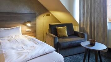 Ein Hotelzimmer mit einem Bett, einem grauen Sessel mit gelbem Kissen, einem kleinen runden Tisch mit einer Tasse und einem gemusterten Vorhang.