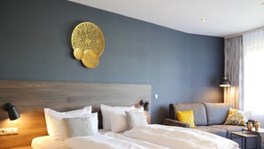 A modern hotel room with a large bed, a sofa, a wall-mounted decorative piece, and a small table with a lamp. - NaturKulturHotel Stumpf (Neunkirchen)