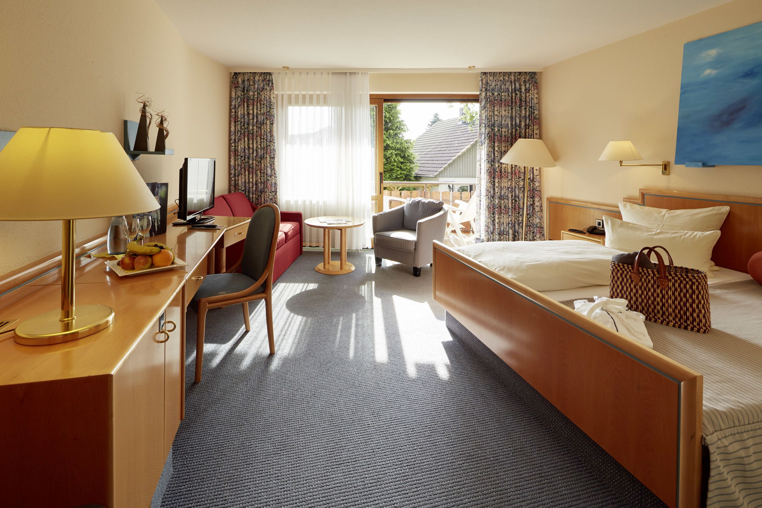 A hotel room with a bed, desk, chair, and a small balcony with curtains.