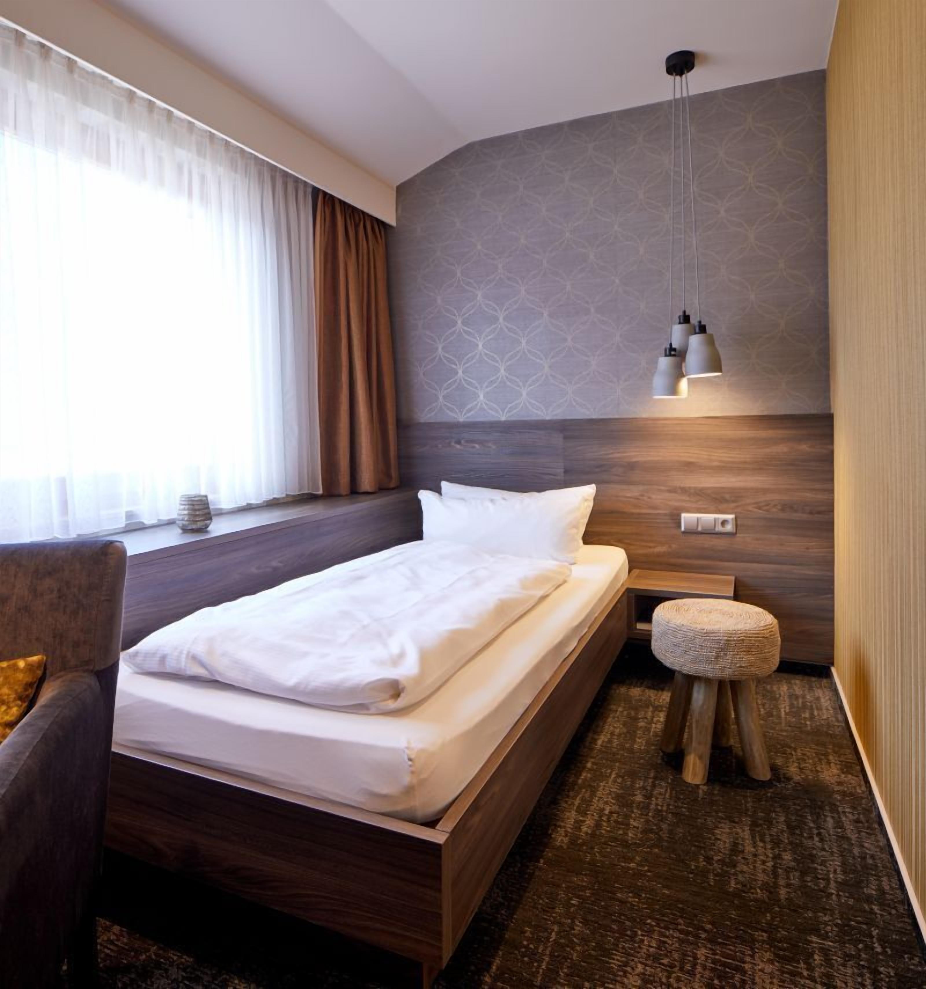 Una habitación de hotel moderna con una cama de madera, un pequeño reposapiés, una mesita de noche y una pared con estampado.