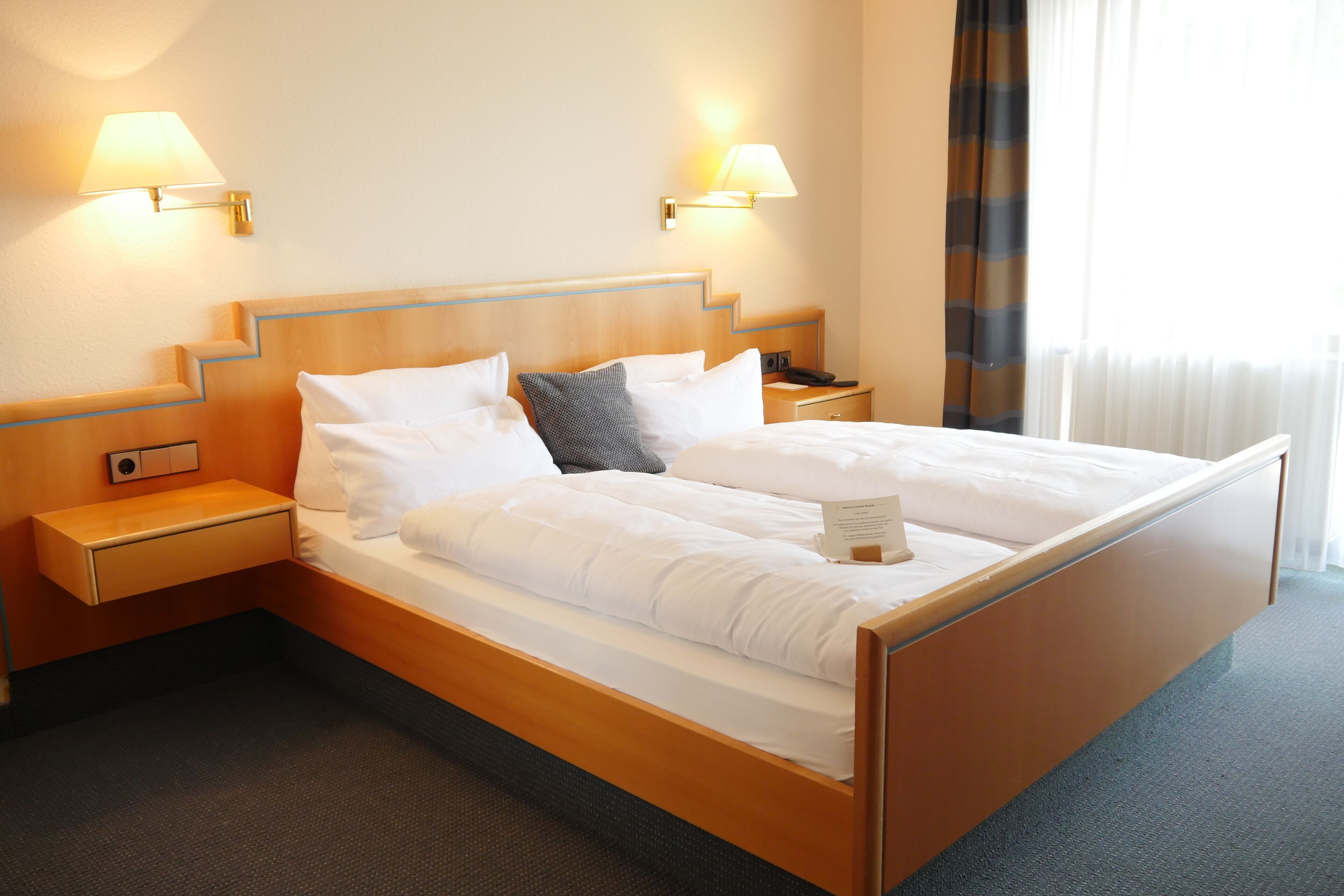 Una cama bien hecha con sábanas blancas, dos lámparas de noche, una mesita de noche y una ventana con cortinas.